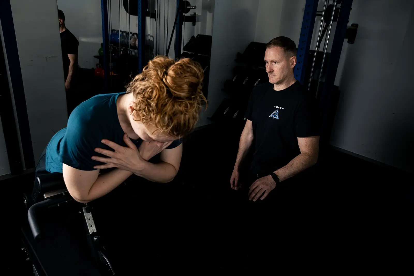 Frau stützt sich auf eine Trainingsbank, Trainer sitzt daneben